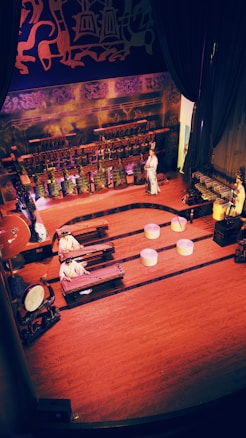 A traditional stage performance with musicians dressed in cultural attire playing various instruments. The setting features ornate decorations and traditional motifs with intricate patterns on the backdrop. Large percussion instruments and string instruments are visible, with performers engaged in a musical presentation.