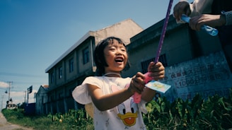 A child joyfully plays outside, holding a toy wand. The scene is set in an urban area with buildings and greenery in the background. Another person is handing the child a bottle of bubble solution.