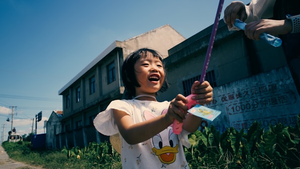 A child joyfully plays outside, holding a toy wand. The scene is set in an urban area with buildings and greenery in the background. Another person is handing the child a bottle of bubble solution.