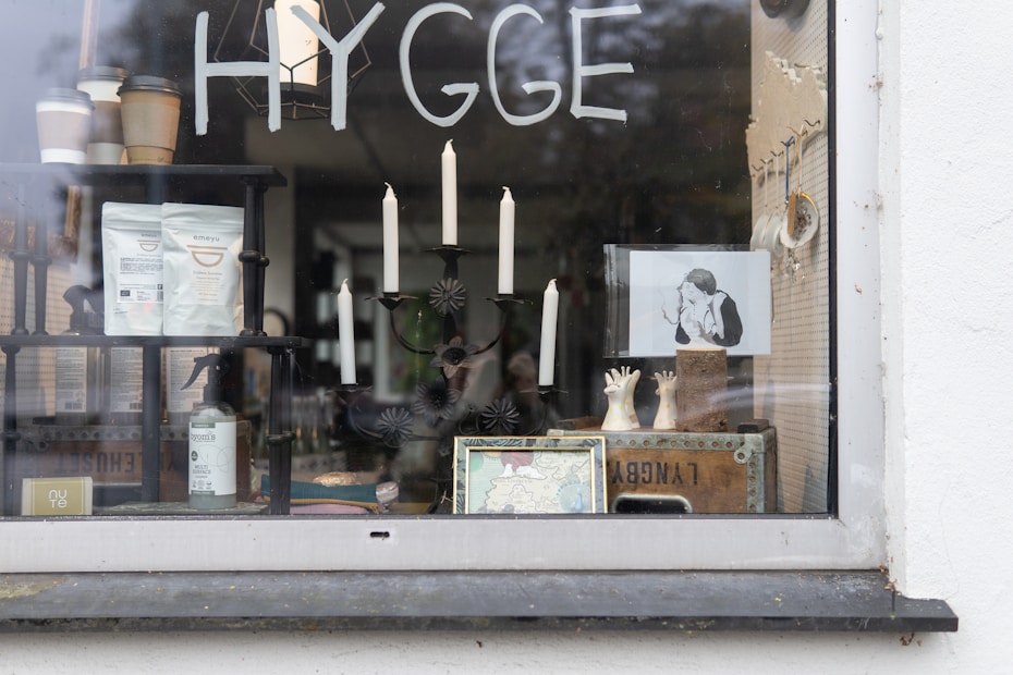 A storefront window displays a variety of items. It features the word 'HYGGE' written on the glass. Inside the window are various objects including a black candlestick holder with white candles, a framed black and white photograph of a person, potted plants, packaged products, a map, and some decorative items such as small sculptures of hands.