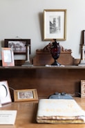 A vintage setup on a wooden surface with multiple framed photographs, some with sepia-toned imagery. An ornate wooden backdrop holds an old ink bottle or lamp in dark red glass. The background features a main framed picture of a historical building or street scene in black and white. Various frames, some empty, are positioned around, and an open book or album with embossed gold details lies in the foreground.