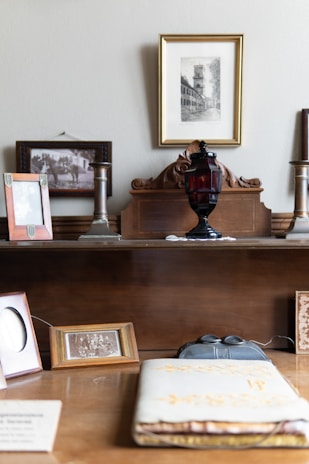 A vintage setup on a wooden surface with multiple framed photographs, some with sepia-toned imagery. An ornate wooden backdrop holds an old ink bottle or lamp in dark red glass. The background features a main framed picture of a historical building or street scene in black and white. Various frames, some empty, are positioned around, and an open book or album with embossed gold details lies in the foreground.