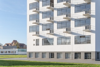 A modernist building with large rectangular windows and white walls, complemented by surrounding green grass and a clear blue sky. The structure has a clean, geometric design with shadows from the window frames casting onto the wall.