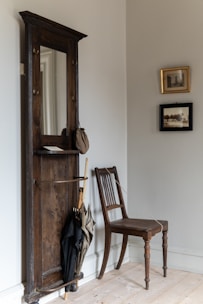 A cozy corner featuring a wooden wall-mounted hall tree with an attached mirror, serving as an umbrella stand holding two umbrellas. A vintage chair with a woven seat sits next to it, and the walls are adorned with two framed black and white photographs.