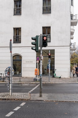 A city street with a modern white building featuring several windows. A storefront labeled 'ANINE BING' is visible, with a display of clothing inside. There is a traffic light showing a red pedestrian signal and a parked e-scooter nearby. The road has marked pedestrian crossings and a round sign covered with stickers. Two people are walking along the sidewalk.