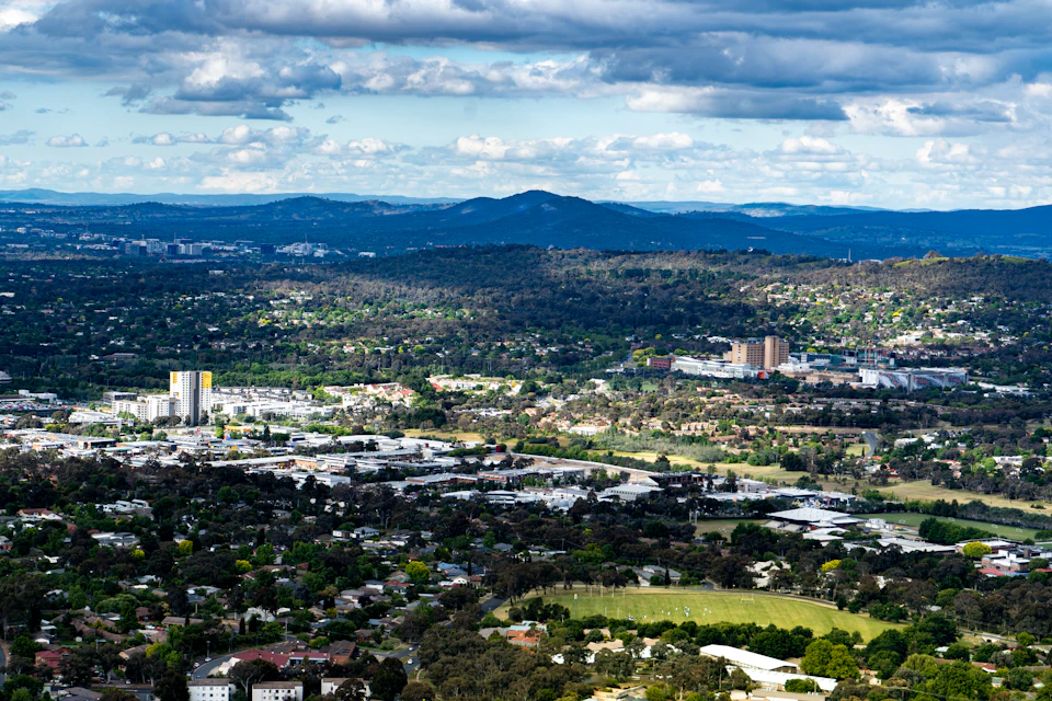 The image presents a panoramic view of a city nestled amidst verdant hills and mountains, with a clear blue sky dotted with fluffy white clouds overhead. The cityscape is a blend of modern skyscrapers and older buildings, creating a harmonious balance between the urban and natural elements. The perspective from above provides an expansive view of the city's layout and its relationship to the surrounding landscape.