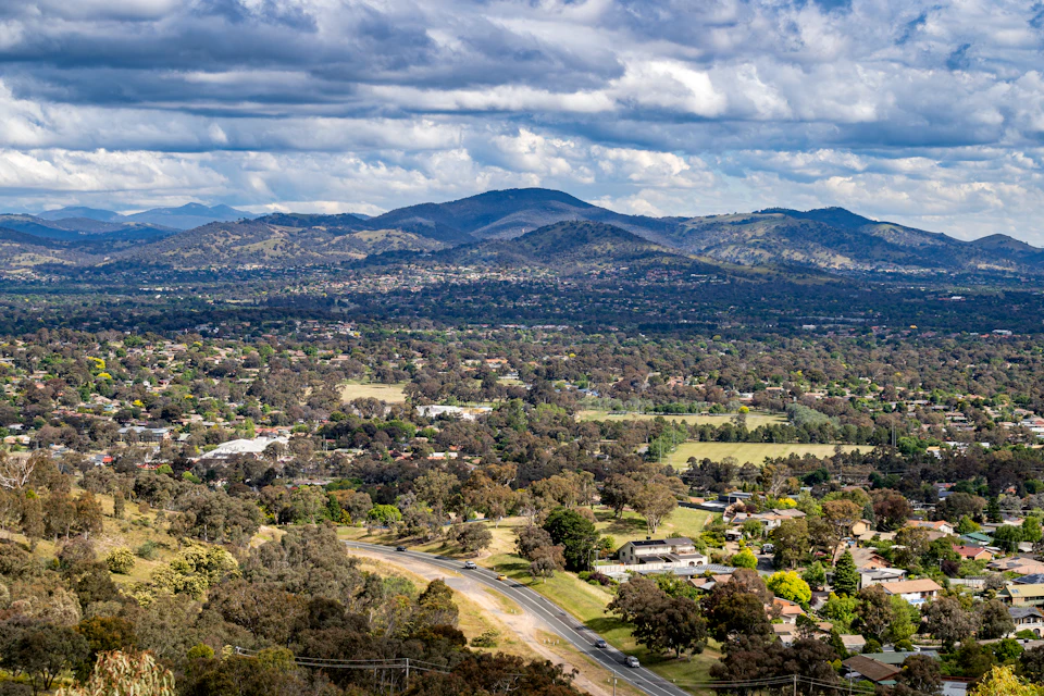 The image presents a panoramic view of a town nestled at the foot of a mountain range, with houses and trees dotting the landscape. The mountains rise majestically in the background, their peaks shrouded in clouds. A winding road cuts through the center of the scene, leading towards the distant horizon where the sky meets the earth. The colors in the image are predominantly green from the vegetation and browns from the buildings and roads.