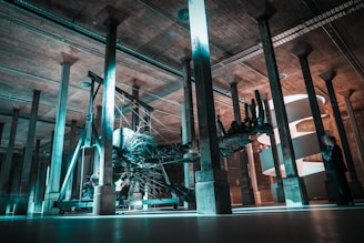 A large, intricate sculpture resembling a tree root structure is suspended in an industrial-looking space supported by numerous concrete columns. The area is dimly lit with striking teal and orange lighting, creating an otherworldly atmosphere. In the background, a helical staircase wraps elegantly behind the sculpture. A man in a suit appears to be examining the installation.