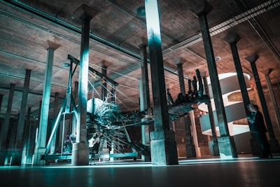 A large, intricate sculpture resembling a tree root structure is suspended in an industrial-looking space supported by numerous concrete columns. The area is dimly lit with striking teal and orange lighting, creating an otherworldly atmosphere. In the background, a helical staircase wraps elegantly behind the sculpture. A man in a suit appears to be examining the installation.