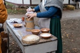 A person dressed in historical or traditional attire is preparing food on a wooden table outdoors. The table is covered with various clay bowls containing ingredients, as well as a wooden container with metal bands. The surrounding area includes fallen leaves and resembles a rustic setting.