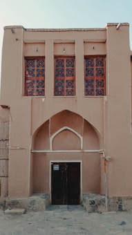 A traditional building with a sand-colored facade features a large, arched doorway at the center, flanked by intricate wooden windows with geometric patterns. The structure is elevated on a stone base and exhibits a minimalist yet historic architectural style.