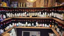 A wooden shelf displays numerous wine bottles, each adorned with a small Santa hat and detailed labels. Above the wine are stacked packages of coffee and various boxes of cookies and chocolates. The arrangement creates a festive and inviting atmosphere.