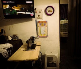 A room with a television mounted on the wall displaying a scene from a movie, a purple framed wall clock, a colorful calendar, a sewing machine on a wooden table with measuring tape, and a white stool. A small air cooler is visible in the partially opened area with a curtain.