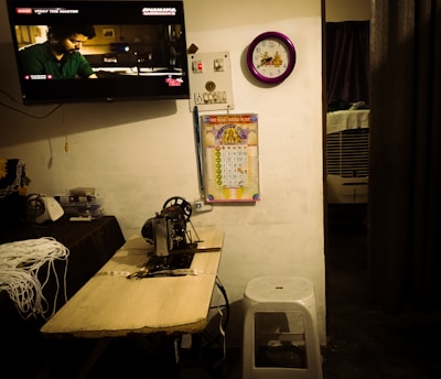 A room with a television mounted on the wall displaying a scene from a movie, a purple framed wall clock, a colorful calendar, a sewing machine on a wooden table with measuring tape, and a white stool. A small air cooler is visible in the partially opened area with a curtain.