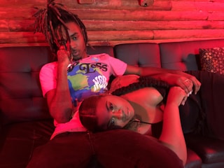A man with dreadlocks and a woman with long braided hair are lounging on a black leather couch. The lighting casts a red hue over the scene, contributing to a relaxed and intimate atmosphere. The man wears a colorful graphic t-shirt, and the woman is in a black outfit.