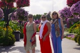 Three women wearing summer dresses and sunglasses are standing on a garden path surrounded by lush greenery and vibrant hanging flower arrangements. The flowers display a range of pink and purple hues, creating a picturesque and cheerful setting.