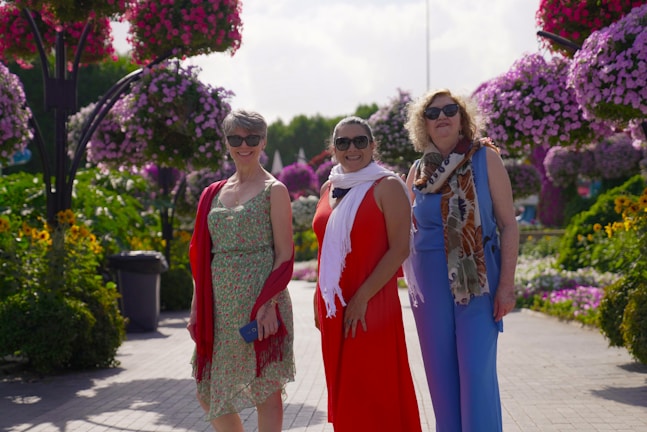 Three women wearing summer dresses and sunglasses are standing on a garden path surrounded by lush greenery and vibrant hanging flower arrangements. The flowers display a range of pink and purple hues, creating a picturesque and cheerful setting.
