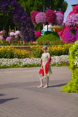 A woman in a flowing dress and delicate scarf, walking through a blooming garden on a bright day.
