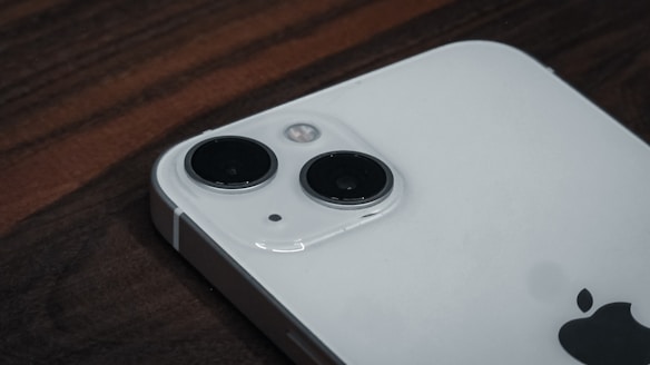 A close-up view of a smartphone lying on a wooden surface, focusing on its dual camera system. The phone is positioned diagonally, showcasing the sleek design and glossy finish with the Apple logo partially visible.