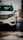 A white car is parked in a garage. The front left side of the vehicle is prominently visible, with distinctive headlights and a recognizable logo on the grill. The background shows some industrial elements like poles and a partial view of the outside through an open gate.