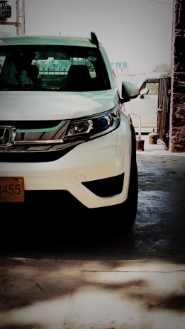 A white car is parked in a garage. The front left side of the vehicle is prominently visible, with distinctive headlights and a recognizable logo on the grill. The background shows some industrial elements like poles and a partial view of the outside through an open gate.