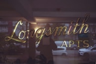 Golden scripted letters spell out 'Longsmith Apts.' on a glass surface, reflecting an urban street scene with parked cars and dim lighting, suggesting a late afternoon or evening setting.