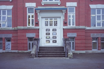 A symmetrical view of a large, classic red brick building with a prominent white doorway at the center. The building features multiple large windows with white trimming. Two short staircases lead up to the main entrance, flanked by concrete railings. The architecture is formal and institutional, possibly indicating a school or government facility.