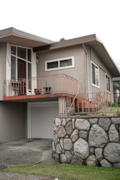 A residential home featuring a mid-century modern architectural style with a light gray facade. The entrance is elevated with a wooden door, sidelight windows, and surrounded by orange-colored metal railings. A garage door is below, alongside a stone wall constructed from large, rounded boulders. The front yard is slightly elevated with a grassy area.