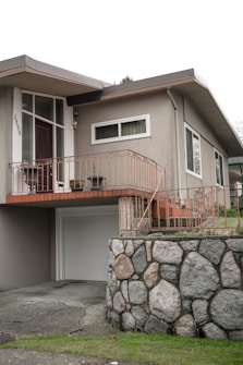 A residential home featuring a mid-century modern architectural style with a light gray facade. The entrance is elevated with a wooden door, sidelight windows, and surrounded by orange-colored metal railings. A garage door is below, alongside a stone wall constructed from large, rounded boulders. The front yard is slightly elevated with a grassy area.