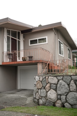 A residential home featuring a mid-century modern architectural style with a light gray facade. The entrance is elevated with a wooden door, sidelight windows, and surrounded by orange-colored metal railings. A garage door is below, alongside a stone wall constructed from large, rounded boulders. The front yard is slightly elevated with a grassy area.