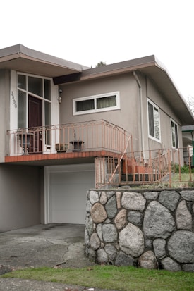 A residential home featuring a mid-century modern architectural style with a light gray facade. The entrance is elevated with a wooden door, sidelight windows, and surrounded by orange-colored metal railings. A garage door is below, alongside a stone wall constructed from large, rounded boulders. The front yard is slightly elevated with a grassy area.