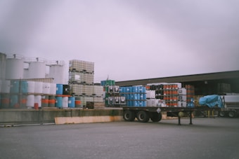 An industrial storage area with stacks of barrels and large containers arranged on pallets. A flatbed trailer is parked nearby, and a part of a cabin or a warehouse is visible in the background. The sky is overcast, creating a dull lighting over the scene.