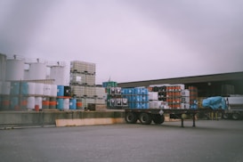 An industrial storage area with stacks of barrels and large containers arranged on pallets. A flatbed trailer is parked nearby, and a part of a cabin or a warehouse is visible in the background. The sky is overcast, creating a dull lighting over the scene.