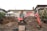 An excavator is positioned in front of a partially excavated yard, which is adjacent to a residential house. The house is situated on a raised foundation with a prominent front door and wide windows. The scene includes piles of dirt and construction materials like concrete blocks and wooden pallets. Shrubbery and other houses are visible on either side.