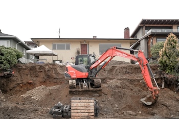 An excavator is positioned in front of a partially excavated yard, which is adjacent to a residential house. The house is situated on a raised foundation with a prominent front door and wide windows. The scene includes piles of dirt and construction materials like concrete blocks and wooden pallets. Shrubbery and other houses are visible on either side.
