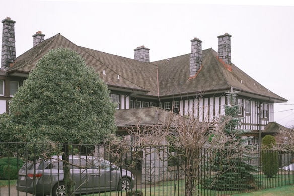 A large, traditional house with four stone chimneys and a steep, shingled roof. The building features timber framing with white walls and large windows. In the foreground, a neatly trimmed bush and a small tree are visible, along with a gray car parked on a paved driveway. The scene is enclosed with a black metal fence, and the surroundings appear to be suburban.