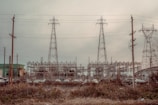 An industrial scene featuring three large electrical pylons standing amidst a complex network of power lines. Surrounding the pylons are multiple poles and transmission equipment, with a backdrop of various industrial structures and buildings. The foreground includes a row of parked vehicles behind a fence and overgrown vegetation.