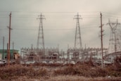 An industrial scene featuring three large electrical pylons standing amidst a complex network of power lines. Surrounding the pylons are multiple poles and transmission equipment, with a backdrop of various industrial structures and buildings. The foreground includes a row of parked vehicles behind a fence and overgrown vegetation.