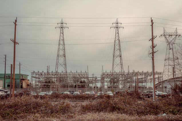 An industrial scene featuring three large electrical pylons standing amidst a complex network of power lines. Surrounding the pylons are multiple poles and transmission equipment, with a backdrop of various industrial structures and buildings. The foreground includes a row of parked vehicles behind a fence and overgrown vegetation.