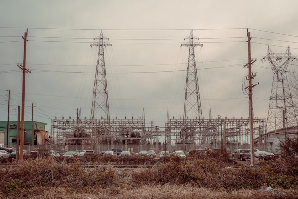 An industrial scene featuring three large electrical pylons standing amidst a complex network of power lines. Surrounding the pylons are multiple poles and transmission equipment, with a backdrop of various industrial structures and buildings. The foreground includes a row of parked vehicles behind a fence and overgrown vegetation.