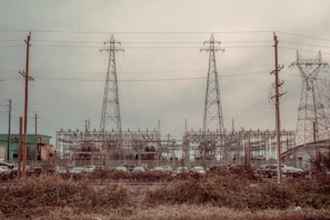 An industrial scene featuring three large electrical pylons standing amidst a complex network of power lines. Surrounding the pylons are multiple poles and transmission equipment, with a backdrop of various industrial structures and buildings. The foreground includes a row of parked vehicles behind a fence and overgrown vegetation.