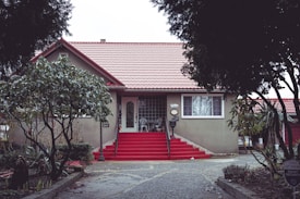 A quaint house with a red-tiled roof and beige exterior is featured, nestled between lush, green trees. The house has a prominent, red-painted staircase leading up to the front door. A security sign is visible on the right side of the image, near the driveway. There are two front windows, and the entrance is sheltered by a small porch roof.