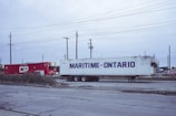 Two shipping containers, one red with 'CP' written on it and another white with 'Maritime-Ontario,' are situated on a gravel surface. Surrounding them are utility poles and wires, suggesting an industrial or logistics area. The sky appears overcast, adding to the subdued atmosphere.