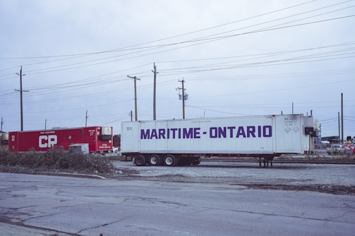 Two shipping containers, one red with 'CP' written on it and another white with 'Maritime-Ontario,' are situated on a gravel surface. Surrounding them are utility poles and wires, suggesting an industrial or logistics area. The sky appears overcast, adding to the subdued atmosphere.