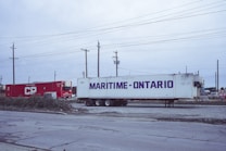 Two shipping containers, one red with 'CP' written on it and another white with 'Maritime-Ontario,' are situated on a gravel surface. Surrounding them are utility poles and wires, suggesting an industrial or logistics area. The sky appears overcast, adding to the subdued atmosphere.