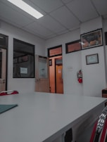 A room with a white ceiling and fluorescent lighting, featuring a large central table and several office doors with glass panels. There are red chairs around the table, framed certificates and art on the walls, and a fire extinguisher mounted on the wall.