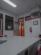 A room with a white ceiling and fluorescent lighting, featuring a large central table and several office doors with glass panels. There are red chairs around the table, framed certificates and art on the walls, and a fire extinguisher mounted on the wall.
