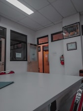 A room with a white ceiling and fluorescent lighting, featuring a large central table and several office doors with glass panels. There are red chairs around the table, framed certificates and art on the walls, and a fire extinguisher mounted on the wall.
