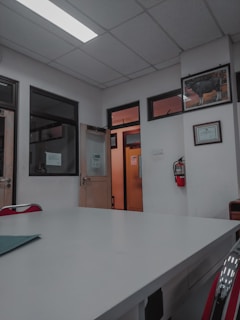 A room with a white ceiling and fluorescent lighting, featuring a large central table and several office doors with glass panels. There are red chairs around the table, framed certificates and art on the walls, and a fire extinguisher mounted on the wall.