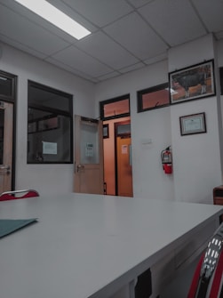 A room with a white ceiling and fluorescent lighting, featuring a large central table and several office doors with glass panels. There are red chairs around the table, framed certificates and art on the walls, and a fire extinguisher mounted on the wall.